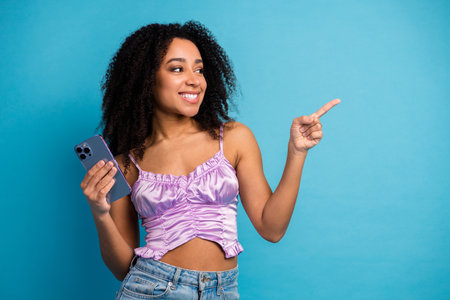 Young stylish girl smiles while pointing and holding a smartphone against a bright blue background in casual chic summer fashionの写真素材
