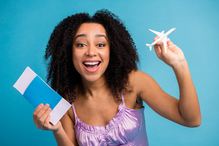 Young woman with curly hair smiling holds a blue ticket and a toy airplane while posing in a purple top against a blue background for travel fashion and lifestyleの写真素材