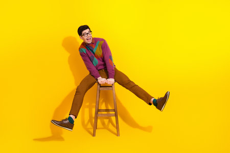 Cheerful young man in colorful attire sitting on a stool against a vibrant yellow background with a joyful expressionの写真素材
