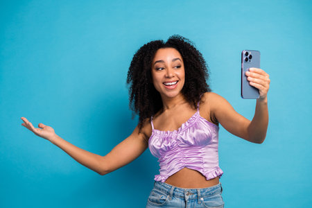 Young woman takes a playful selfie with a smartphone against a bright blue background in stylish...