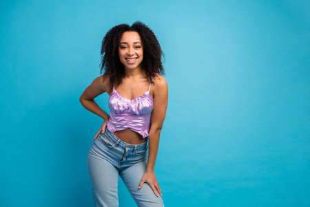 Young woman with curly hair wearing a lilac top posing against a blue studio backdrop with a stylish...