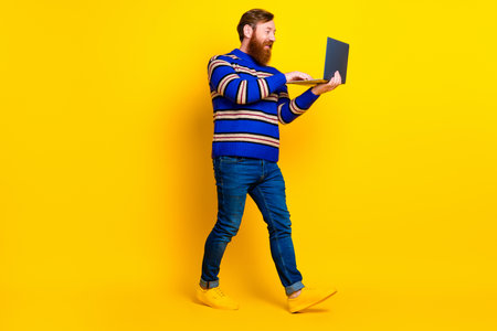 Vibrant man walking confidently in a striped sweater with a laptop against a bright yellow background embodying creativity and energyの写真素材