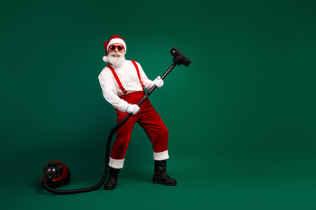 Cheerful Santa Claus in red costume cleaning with a vacuum cleaner on a green background celebrating the holiday seasonの写真素材