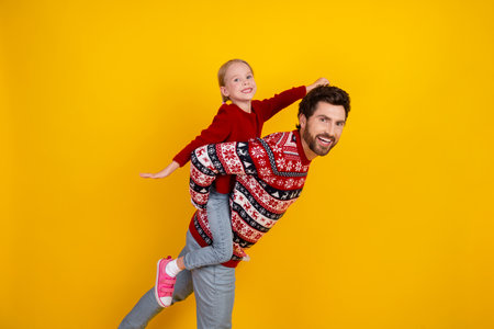Father giving a playful piggyback ride to his smiling daughter on a vibrant yellow background during a joyful winter dayの写真素材