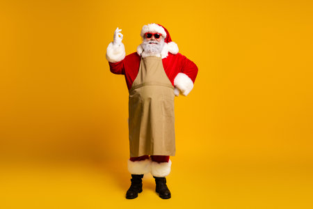 Santa Claus in apron poses with an okay gesture on yellow background, wearing sunglasses and showcasing festive cheerの写真素材