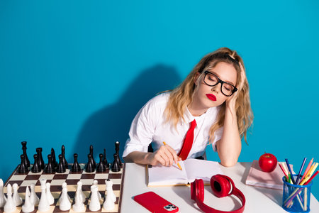Young female student in stylish uniform with blonde hair writing on a notepad while sitting against...