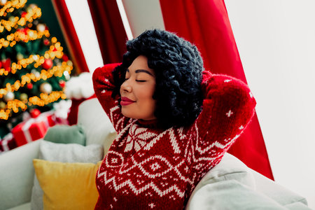 Cozy smiling woman in a red sweater relaxing on the couch during the holiday season with festive Christmas decorationsの写真素材