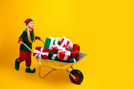 Festive young elf pushing a wheelbarrow filled with holiday gifts against a vibrant yellow backdropの写真素材