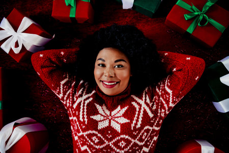 Festive woman in a cozy red sweater smiles surrounded by wrapped Christmas presents in a warm...