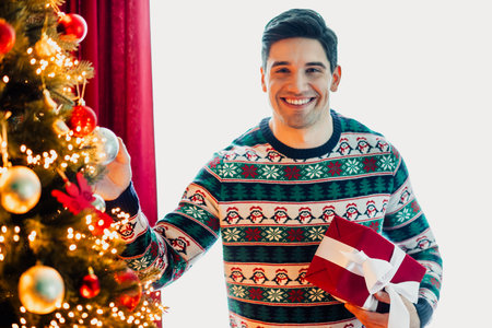 Young man decorating Christmas tree and holding present, enjoying festive holiday atmosphere indoors