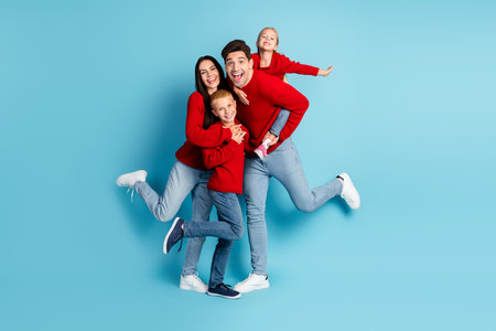 Cheerful family jumping together in festive red sweaters against a bright blue backdrop for a holiday celebration.の写真素材