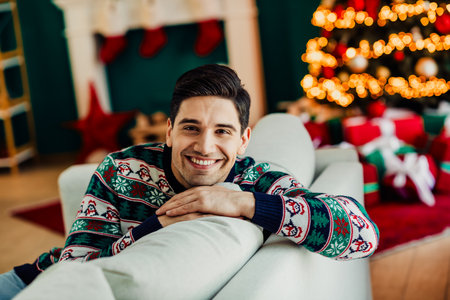 Young man in festive sweater smiling cheerfully surrounded by Christmas decorations in cozy home settingの写真素材