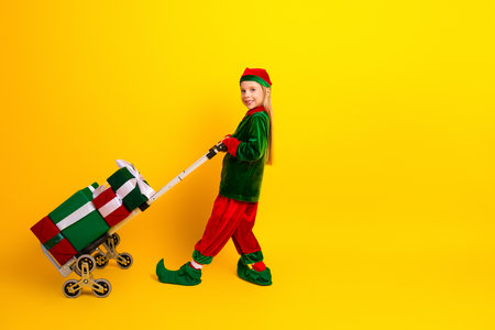 Smiling little girl dressed as a festive Christmas elf pushing a trolley of gifts against a bright yellow backgroundの写真素材