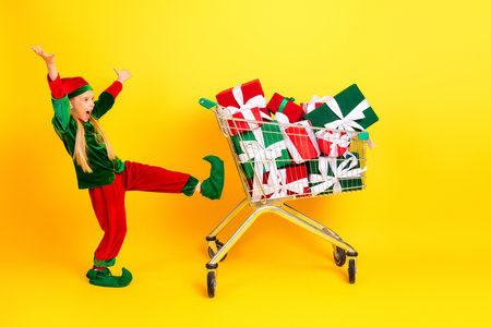 Happy child in elf costume posing energetically with gift-filled cart against bright background...