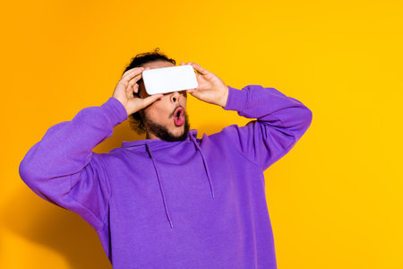 Young man in a purple hoodie interacting with smartphone against a yellow background showing...