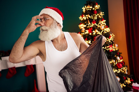Elderly man with Santa hat holding trash bag in a decorated room with glowing Christmas tree, smiling intimatelyの写真素材