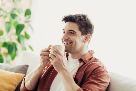 Relaxed young man enjoying leisure time at home with a cup of coffee, sitting on a cozy couch on a bright dayの写真素材