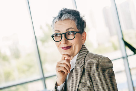 Confident senior businesswoman with glasses in a stylish jacket thinking in a modern office...