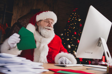 Santa at his desk greets with a green card in a cozy home office while a Christmas tree glows softly and festive lights surround the scene for a warm holiday momentの写真素材