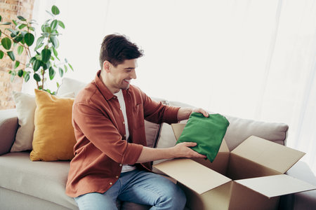 Young man happily unpacking a box at home in a cozy living room featuring a sofa and open windows
