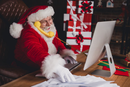 Santa at home workshop talking on a bright yellow phone while organizing gifts beside a glowing christmas tree in a cozy desk setupの写真素材