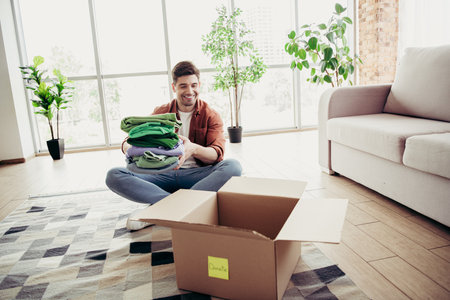 Young man happily organizes clothes for donation at home, sitting on the carpet in a bright modern...