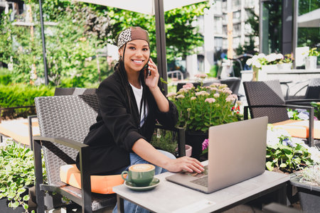 Playful urban woman at sunny cafe working on laptop chatting on phone surrounded by plants and city vibesの写真素材