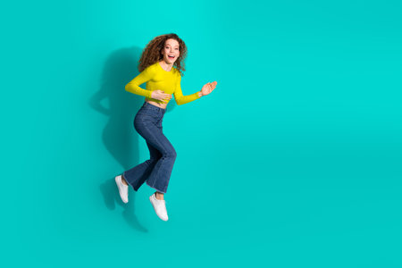 Charming young girl in stylish casual attire jumping joyfully against a vibrant teal background...