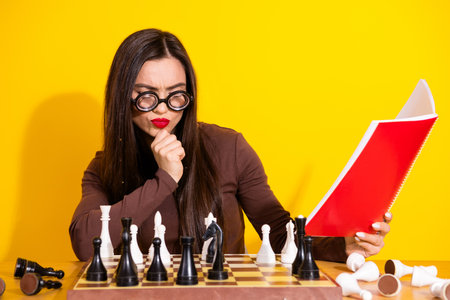 Young woman concentrating on chess game while reading notes in bright yellow background scene