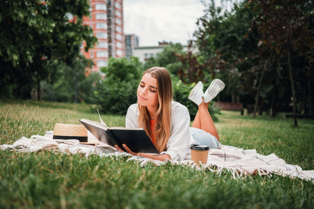 Young woman relaxing in a city park reads a book on a blanket with coffee as tall buildings and green trees frame a calm sunny summer lifestyle stock photo sceneの写真素材