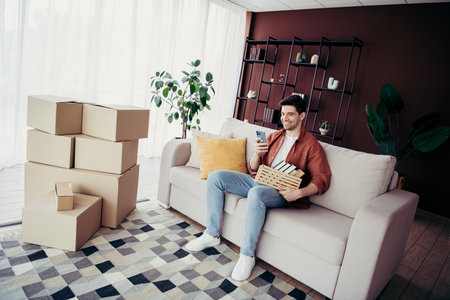 Young man relaxing on couch unpacking books in modern apartment during moving day with packed boxes...