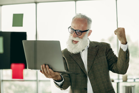 Elderly businessman celebrating success with laptop in office showcasing confidence and eleganceの写真素材