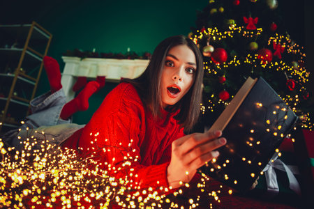 A cheerful woman in a red sweater finds a playful surprise under a twinkling Christmas tree at home for a cozy festive momentの写真素材