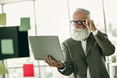 Mature businessman with a white beard in stylish attire working on a laptop in a modern office settingの写真素材