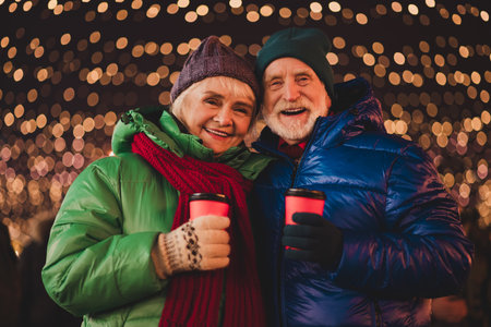 Funny warm grandparents couple enjoy hot drinks under festive lights outdoors together in a busy city during christmas seasonの写真素材