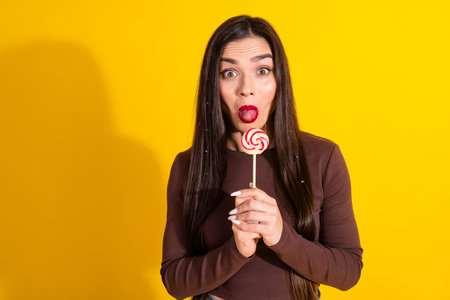 Cute young woman enjoying a lollipop with a vibrant yellow background for joyful lifestyle and...