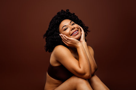 Smiling confident woman with natural curly hair posing against brown background showcasing self-confidence and positivityの写真素材