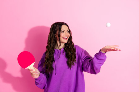 Young woman in casual sweatshirt holding a ping pong paddle, posing with a smile against a vibrant pink backdropの写真素材