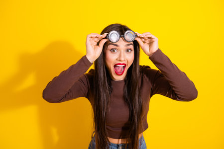 Happy young woman in a stylish brown top with goggles on a vibrant yellow background expressing...