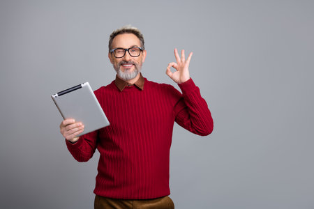 Smiling businessman in a red sweater holding a tablet while making an OK gesture with a confident expressionの写真素材
