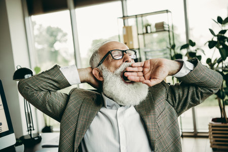 Elderly businessman yawning and reflecting in an office setting, demonstrating tiredness yet professionalism and styleの写真素材