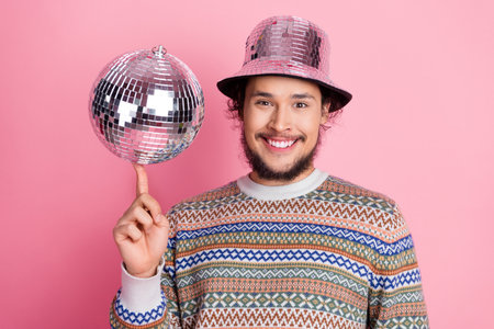 Smiling man wearing a stylish sweater and mirrored hat against a pink background, holding a silver disco ball on his fingerの写真素材