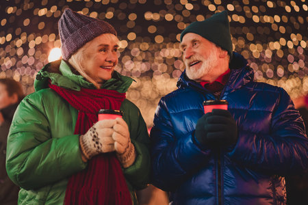 A warm moment as a couple and grandparents share coffee under festive lights during a winter night in the city while wrapped in colorful jacketsの写真素材