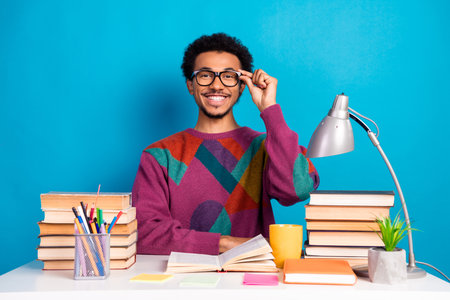 Funny stylish man with glasses studying at desk surrounded by books and supplies against bright blue...