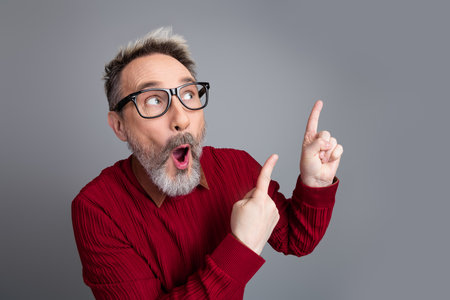 Mature businessman in red sweater showcasing surprise and excitement while pointing with both hands on a gray backgroundの写真素材