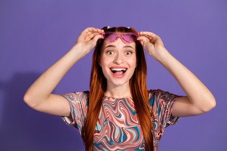 Cheerful young woman with colorful top holding pink glasses on vibrant purple background showing...