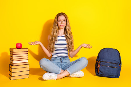 Young female student sitting casually with confused expression among book and a backpack on a yellow backgroundの写真素材