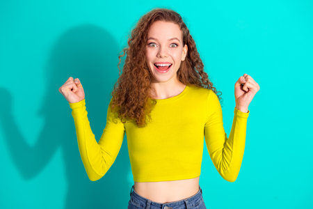 Charming cheerful woman with curly red hair celebrates in casual outfit against a bright teal...