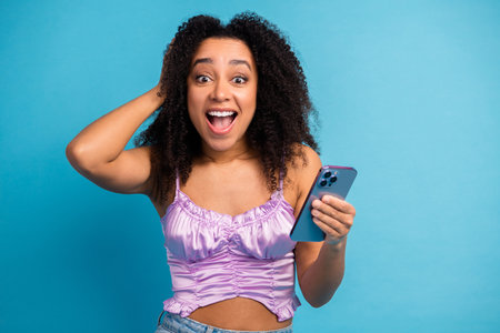Young woman with curly hair smiles at her phone against a blue background showcasing casual chic...