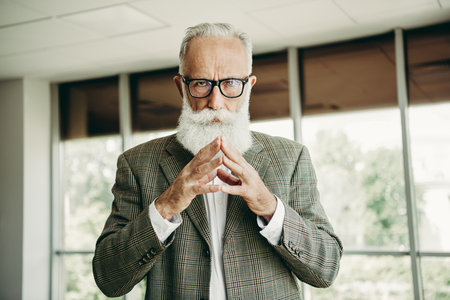 Confident senior professional businessman in formal attire with a stylish white beard in a modern office interiorの写真素材